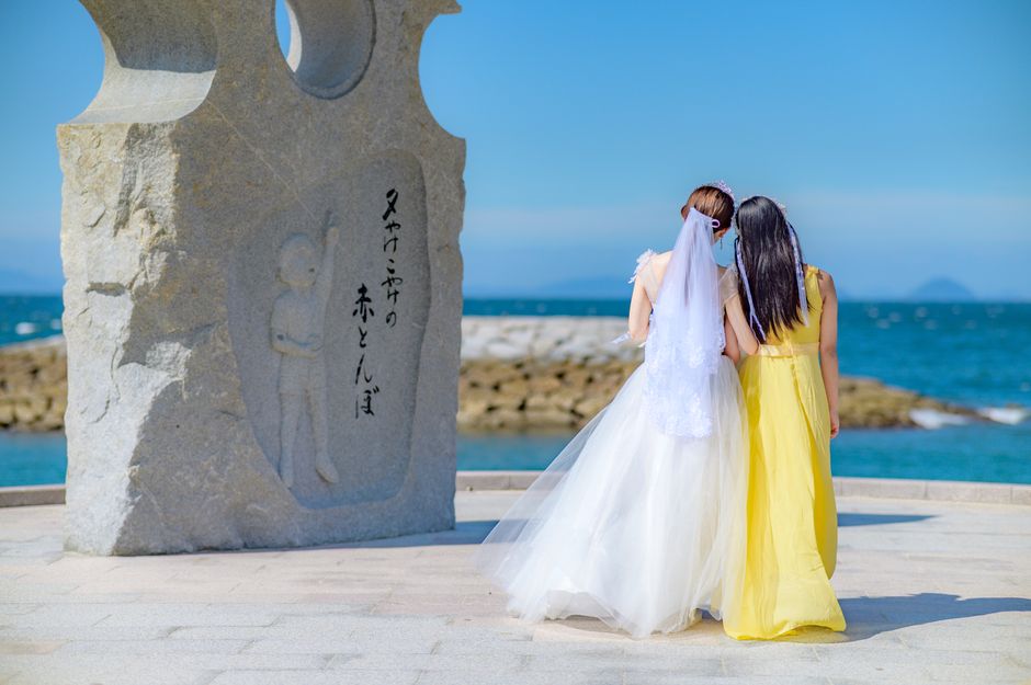 海辺の記念碑のそばで、ウェディングドレスを着た花嫁と黄色いドレスを着た友人。